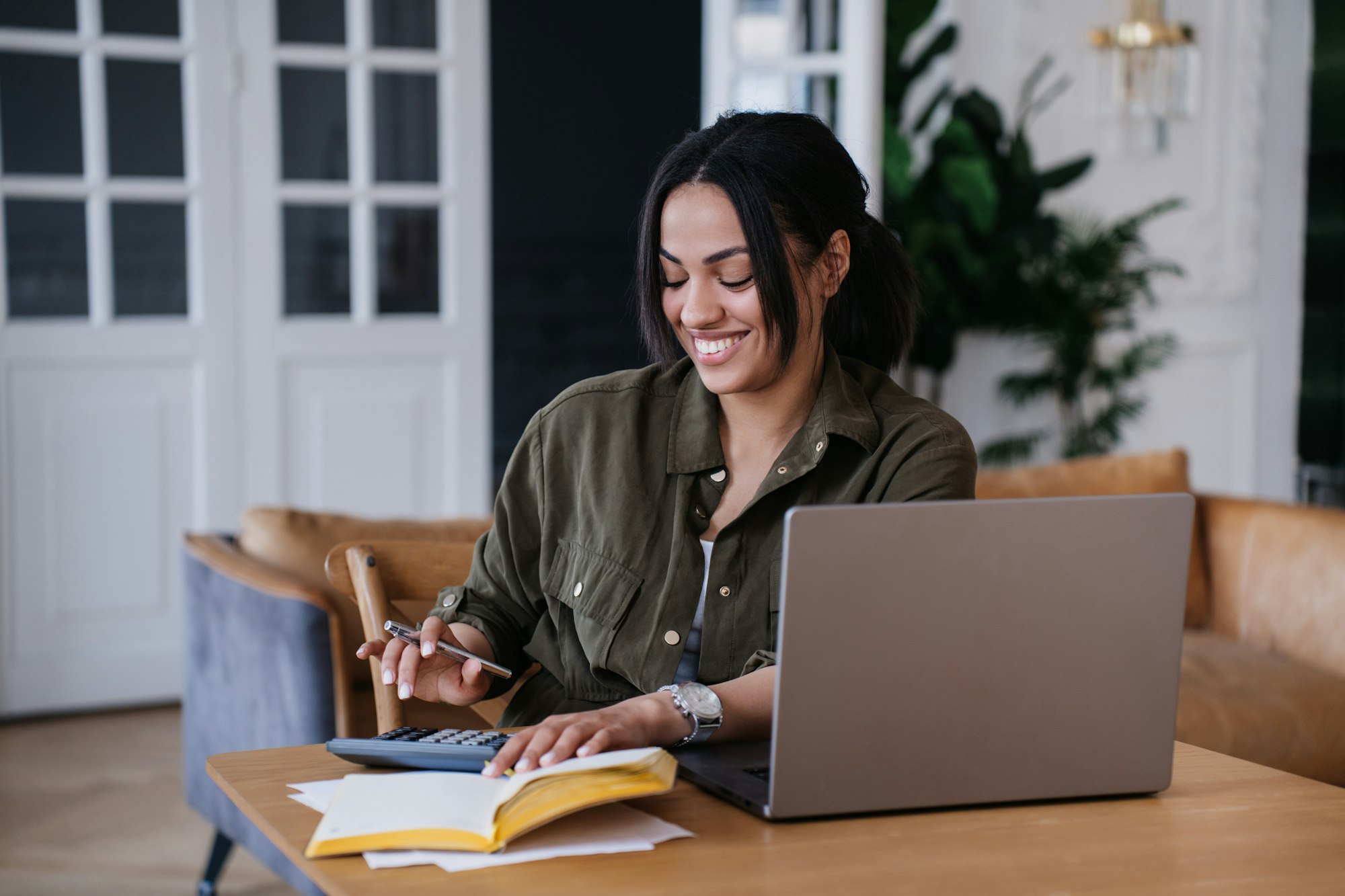 Cheerful accountant using calculator sits at desk with laptop, smiles broadly, enjoying work home